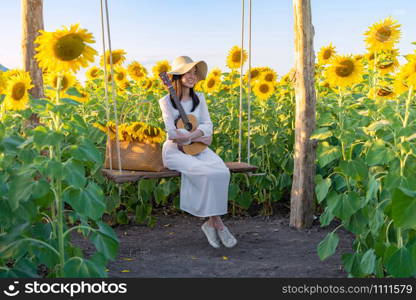 An Asian woman enjoying, relaxing, playing an ukulele in sunflower field on swing during travel holidays vacation trip outdoors at natural garden park at sunset in Lopburi, Thailand