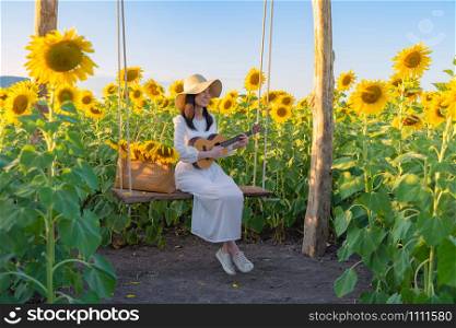 An Asian woman enjoying, relaxing, playing an ukulele in sunflower field on swing during travel holidays vacation trip outdoors at natural garden park at sunset in Lopburi, Thailand