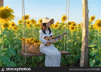 An Asian woman enjoying, relaxing, playing an ukulele in sunflower field on swing during travel holidays vacation trip outdoors at natural garden park at sunset in Lopburi, Thailand