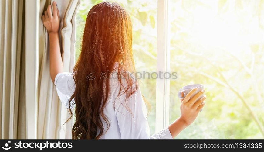 An Asian woman stood holding a coffee cup and opened the curtain on the window after waking up in the morning of the day at home