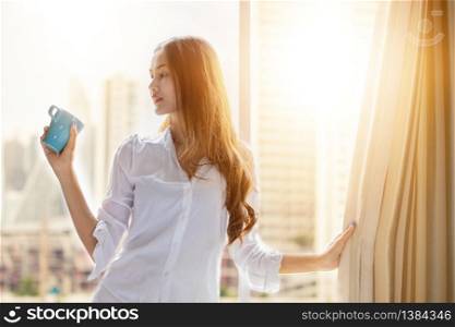 An Asian woman stood holding a coffee cup and opened the curtain on the window after waking up in the morning of the day at home