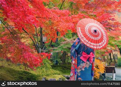 An Asian woman wearing Japanese traditional kimono with red umbrella standing with Red maple leaves or fall foliage in Autumn season during travel holidays vacation trip outdoors in Kyoto City, Japan.