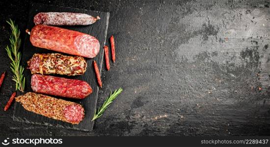 An assortment of delicious salami sausage on a stone board with rosemary. On a black background. High quality photo. An assortment of delicious salami sausage on a stone board with rosemary.