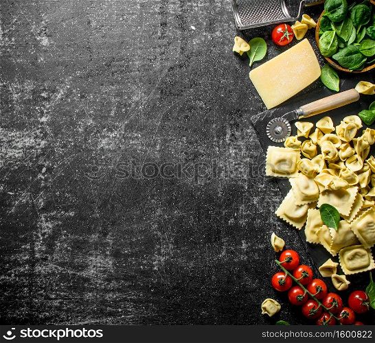 An assortment of different types of raw pasta with cheese, tomatoes and herbs. On dark rustic background. An assortment of different types of raw pasta with cheese, tomatoes and herbs.