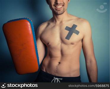 An athletic young shirtless man is holding pads and is ready for some muay thai or mixed martial arts training