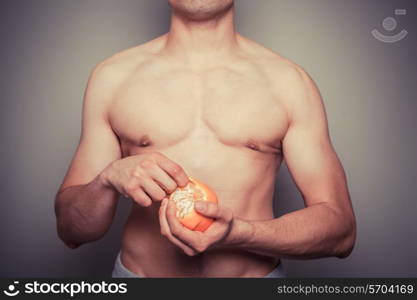 An athletic young shirtless man is peeling an orange