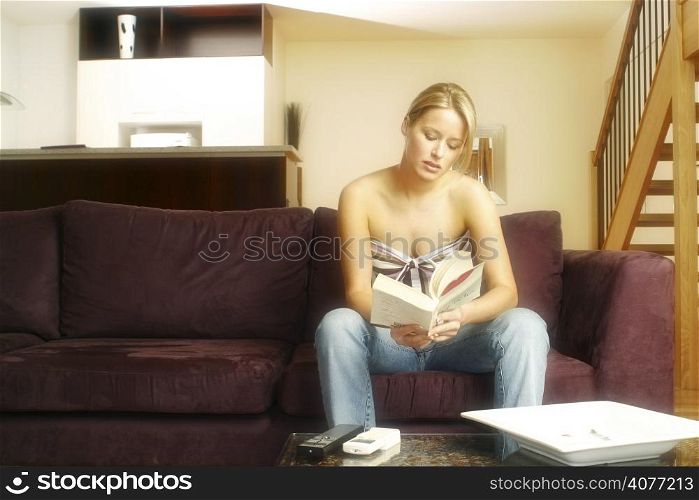 An attractive blond woman relaxes on the couch reading a book.