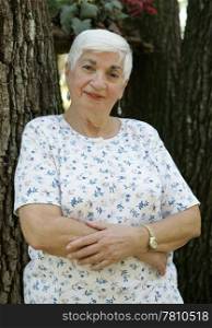 An attractive, elderly grandmother leaning against a tree with her arms folded.
