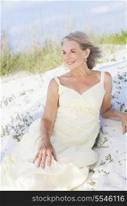 An attractive elegant classy senior woman in a yellow sun dress sitting on a white sand beach with grass and a blue sky behind her.