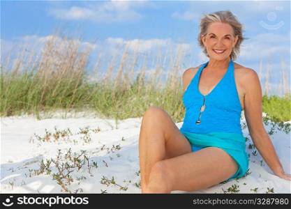 An attractive elegant senior woman in a blue swimming costume sitting on a white sand beach with grass and a blue sky behind her.