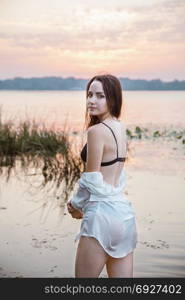 an attractive girl in lingerie and a white shirt is standing by the river at dawn. beauty, fashion, portrait.