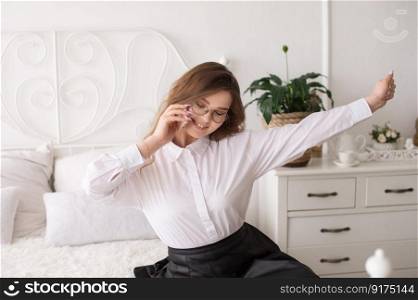An attractive girl works as a remote worker in her apartment. Scandinavian bedroom interior. Remote work.. An excited successful European woman with a phone raises her hands. A remote worker in her apartment.