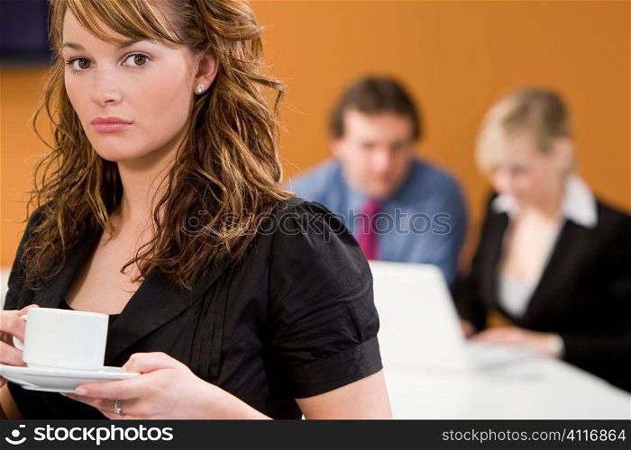 An attractive young female executive takes a coffee break while her colleagues work on a laptop behind her