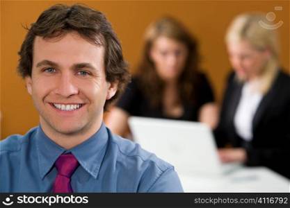 An attractive young male executive in focus in the foreground while his female colleagues work on a laptop behind her