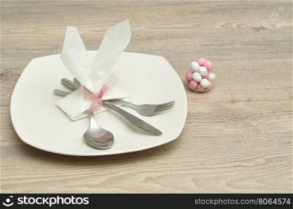 An Easter place setting with the napkin in the shape of bunny ears displayed with a plate, knife, fork, spoon and a bowl of candy