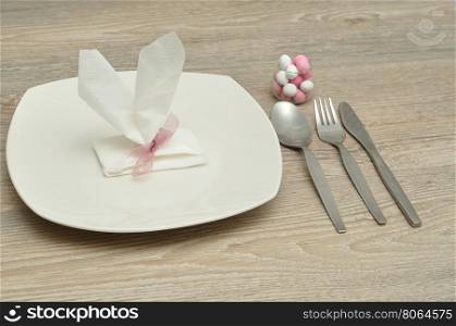 An Easter place setting with the napkin in the shape of bunny ears displayed with a plate, knife, fork, spoon and a bowl of candy