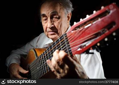 An elderly man in white shirt playing an acoustic guitar. Dark background.
