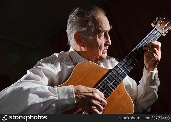 An elderly man in white shirt playing an acoustic guitar. Dark background.