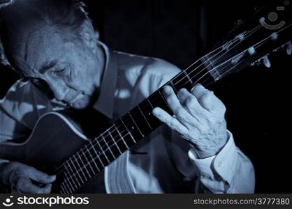 An elderly man in white shirt playing an acoustic guitar. Dark background. Monochrome. Focus on the hand.