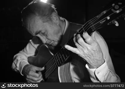 An elderly man in white shirt playing an acoustic guitar. Dark background. Monochrome. Focus on the hand.