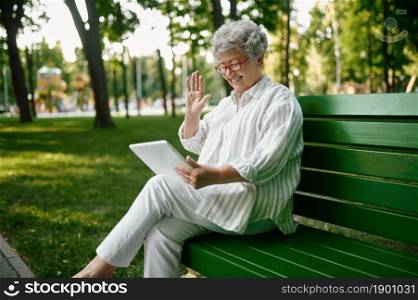 An elderly woman in glasses using laptop on the bench in summer park. Aged people lifestyle. Pretty grandmother having fun outdoors, old female person on nature. An elderly woman in glasses using laptop on bench