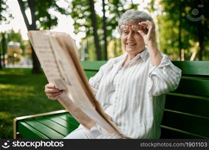 An elderly woman reading newspaper on the bench in summer park. Aged people lifestyle. Pretty grandmother having fun outdoors, old female person outdoors. An elderly woman reading newspaper on the bench
