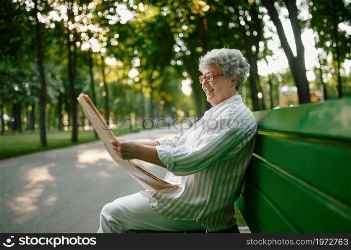 An elderly woman reading newspaper on the bench in summer park. Aged people lifestyle. Pretty grandmother having fun outdoors, old female person on nature. An elderly woman reading newspaper on the bench