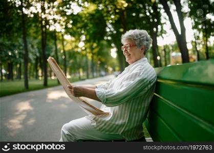 An elderly woman reading newspaper on the bench in summer park. Aged people lifestyle. Pretty grandmother having fun outdoors, old female person on nature. An elderly woman reading newspaper on the bench