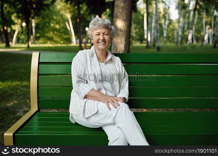 An elderly woman sitting on the bench in summer park. Aged people lifestyle. Pretty grandmother having fun outdoors, old female person on nature. An elderly woman sitting on bench in summer park