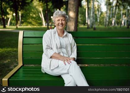 An elderly woman sitting on the bench in summer park. Aged people lifestyle. Pretty grandmother having fun outdoors, old female person on nature. An elderly woman sitting on bench in summer park