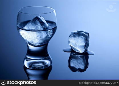 An elegant shot glass with vodka and ice along with a melting ice cube on reflective surface.