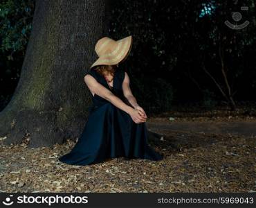 An elegant woman wearing a dress and a hat is relaxing under a tree in the park