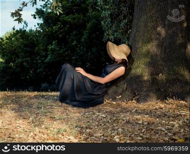 An elegant woman wearing a dress and a hat is relaxing under a tree in the park