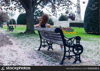 An elegant young woman is sitting on a park bench with cherry blossom on the ground