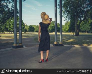 An elegant young woman wearing a hat and a dress is standing in a bandstand in a park at sunset