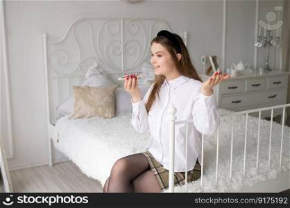 An emotional girl with a red manicure is sitting on the bed in the room and recording a voice message. A cute girl with a red manicure is sitting on the bed in the room and recording a voice message