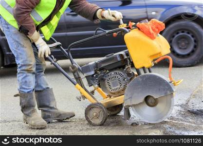 An employee of a road maintenance service in a green reflective vest removes old asphalt with a gasoline cutter during repairs on the roadway.. A worker in a green reflective vest cuts a piece of bad asphalt with a gasoline cutter on the carriageway.