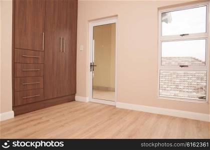 An empty bedroom of a new home with dark brown wooden cupboards, light brown laminated floor, aliminium frame winows and glass door to the patio, as viewed from inside the room.
