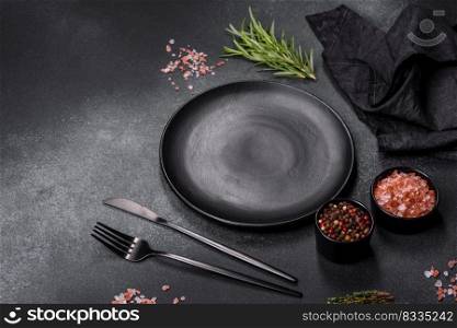 An empty black plate on a dark concrete background with spices, herbs and cutlery. Home dinner cooking. An empty black plate on a dark concrete background with spices and herbs