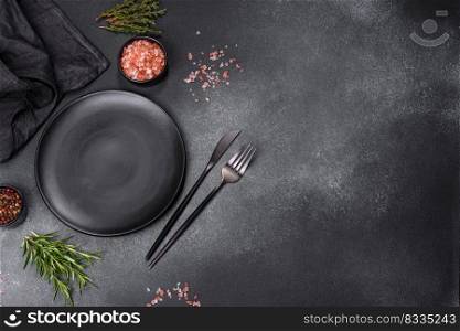 An empty black plate on a dark concrete background with spices, herbs and cutlery. Home dinner cooking. An empty black plate on a dark concrete background with spices and herbs