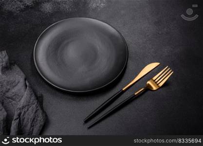 An empty black plate on a dark concrete background with spices, herbs and cutlery. Home dinner cooking. An empty black plate on a dark concrete background with spices, herbs and cutlery