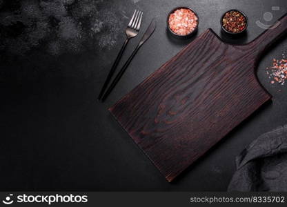 An empty black plate on a dark concrete background with spices, herbs and cutlery. Home dinner cooking. An empty black plate on a dark concrete background with spices, herbs and cutlery