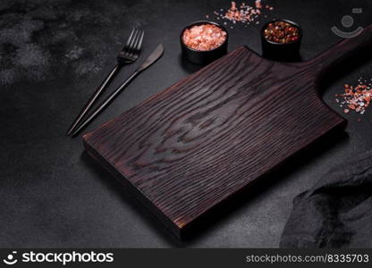 An empty black plate on a dark concrete background with spices, herbs and cutlery. Home dinner cooking. An empty black plate on a dark concrete background with spices, herbs and cutlery