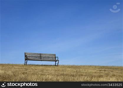 An empty park bench made of wood on dry grass is outlined against a blue sky with light clouds.