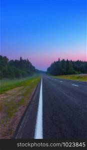 An empty straight asphalt road leads to a pink dawn under the cloudless blue sky in the misty summer morning. Russian federal highway A119. Selective soft focus, space for copy.