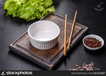 An empty white bowl and sticks on a wooden cutting board on a dark concrete background. Asian food. An empty white bowl and sticks on a wooden cutting board