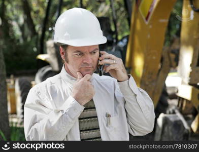 An engineer on a construction site talking on his phone and thinking.