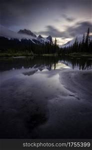 An evening in the Bow Valley as the mountains of Canmore reflect in a creek with the setting crescent moon above.