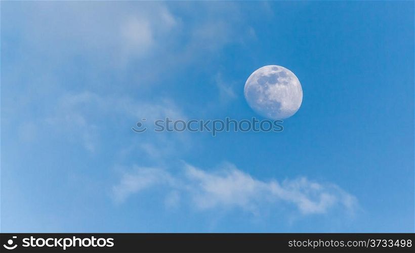 An image of the moon during the day with a clear blue sky and scattered clouds in the background