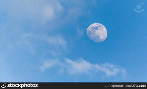 An image of the moon during the day with a clear blue sky and scattered clouds in the background